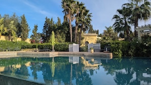 A pool with clear water reflecting surrounding trees and a building. There are lounge chairs and a white fence with lattice panels. - Les Cypres Hotel Restaurant  (Moulay Abdallah)