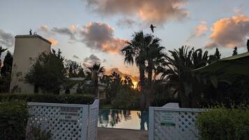 Ein Poolbereich mit einem Tor, im Hintergrund ein Sonnenuntergang, umgeben von Palmen und einem Gebäude.