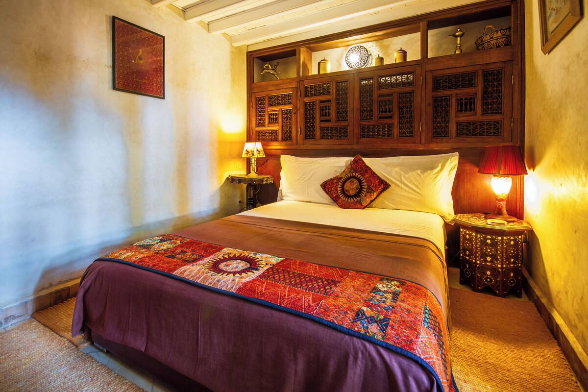 A room with a bed, a nightstand, a wall-mounted shelf with decorative items, and a framed picture on the wall.