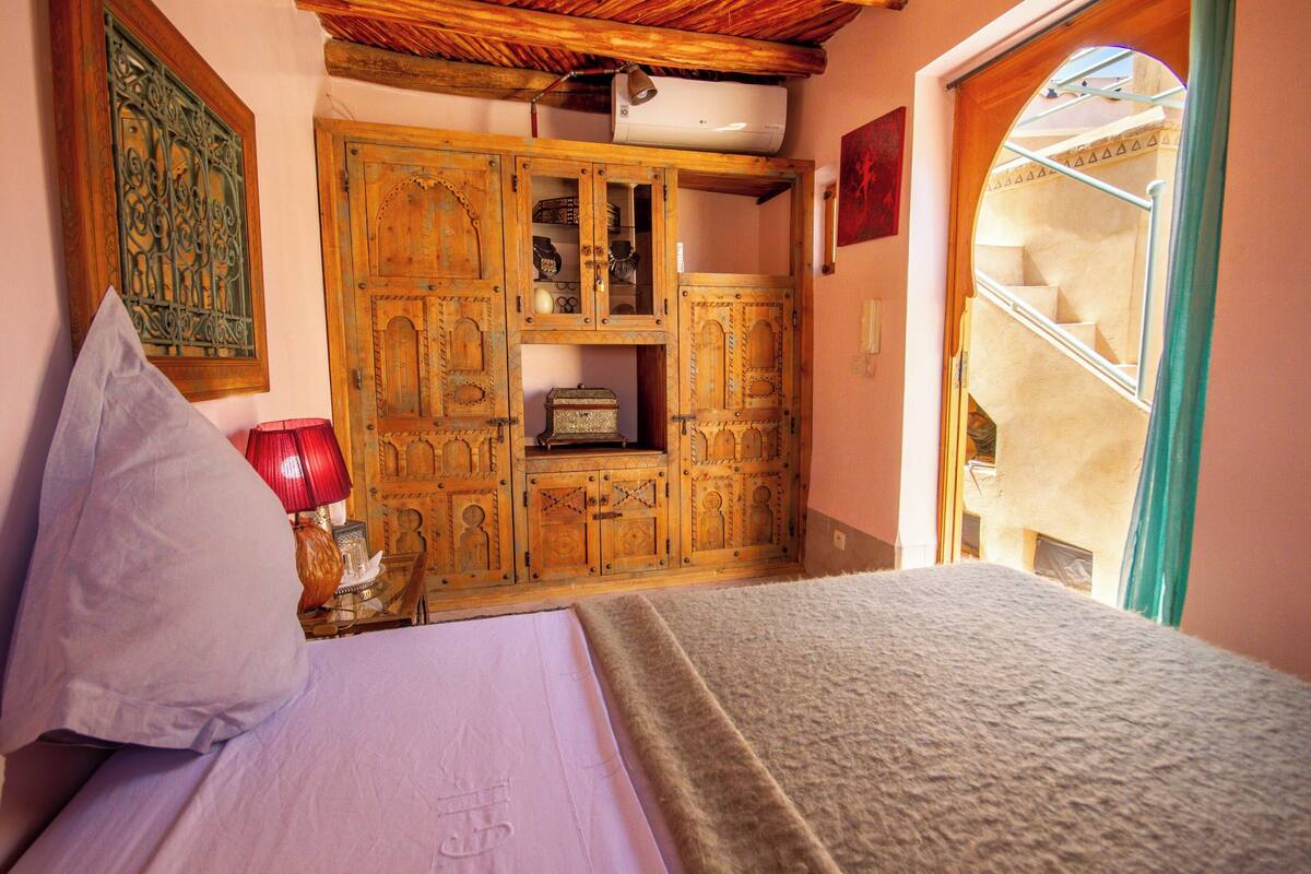 A bedroom with a bed, a wooden wardrobe, a red lamp, and a window with a view of a staircase.
