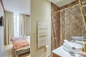 Una habitación de hotel moderna con cama, calefacción integrada, cabina de ducha de mármol y baño con lavamanos y dispensador de toallas.