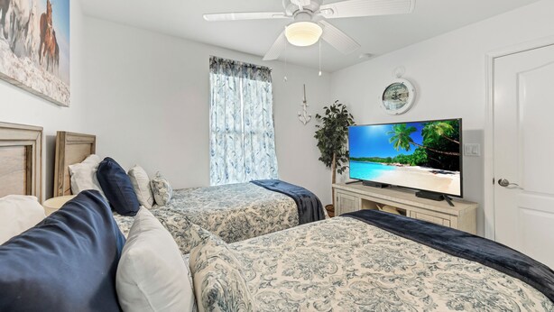 A bedroom with two beds, a ceiling fan, a TV displaying a beach scene, a clock, and a window with curtains. - Spacious beachfront condo with community pool, located on the first floor of the (Port Aransas)