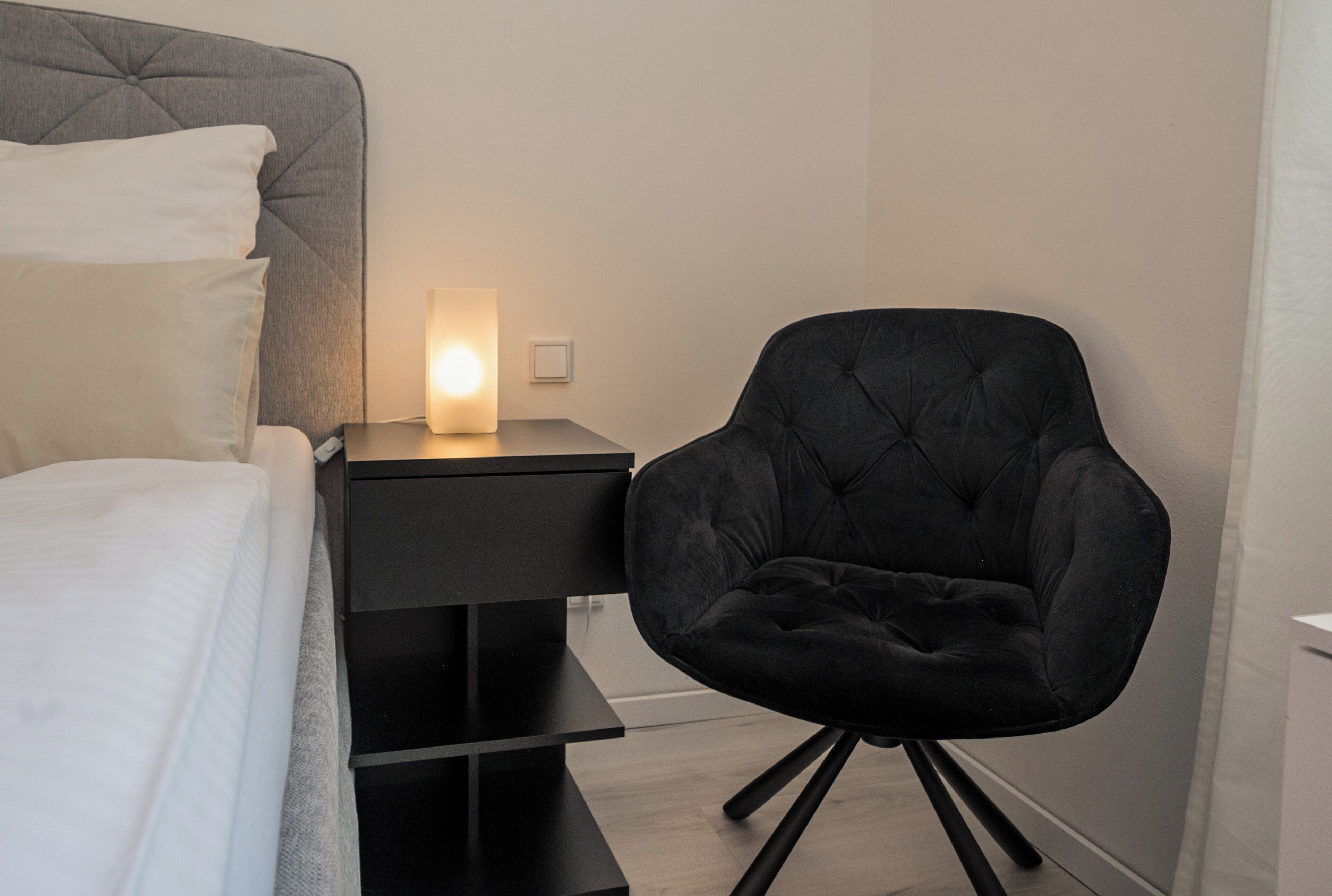 Ein Schlafzimmer mit einem Bett, einem Nachttisch mit Lampe und einem schwarzen Polsterstuhl.