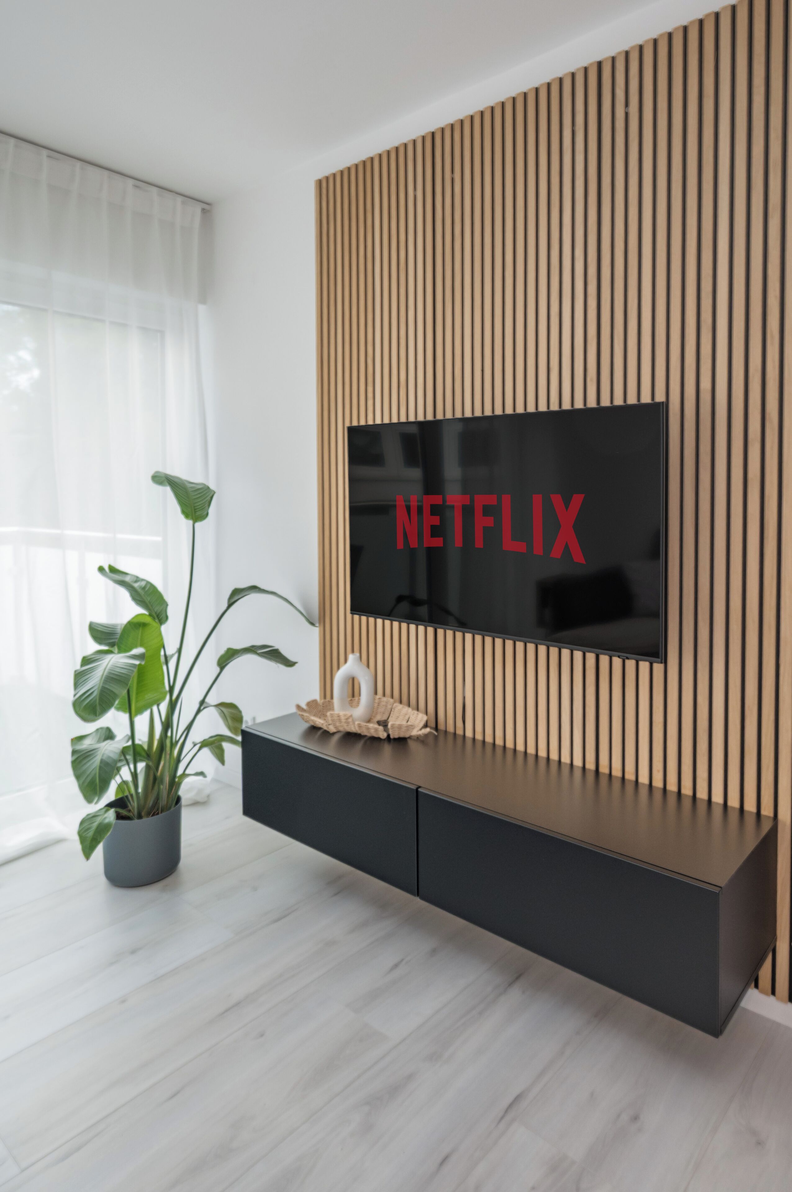 Ein modernes Wohnzimmer mit einem an einer Holzpaneelwand befestigten Flachbildfernseher, einer schwarzen Unterhaltungseinheit darunter und einer Zimmerpflanze links.