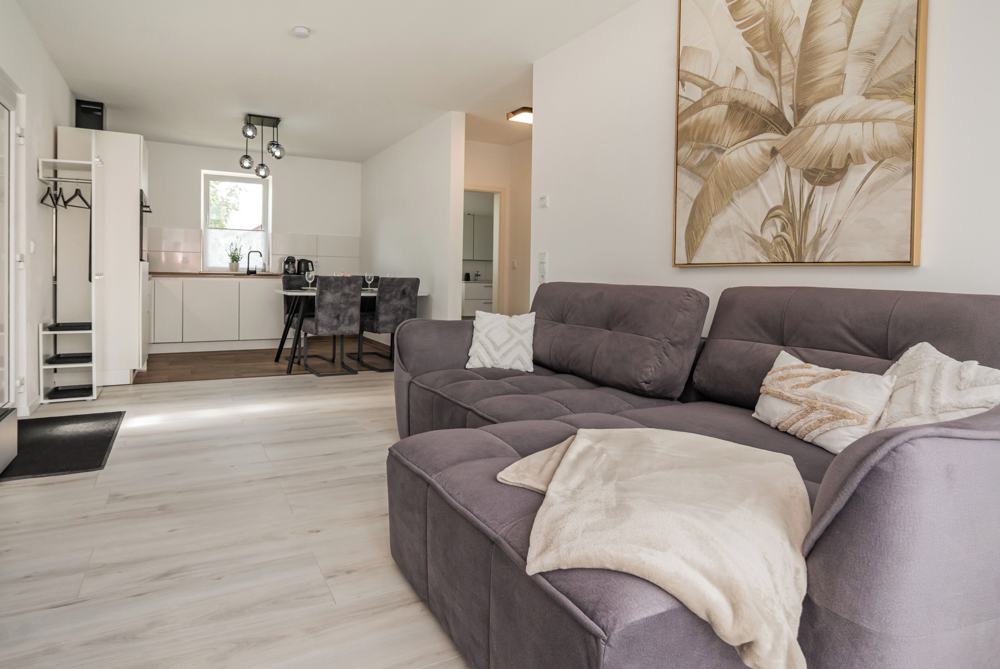 A modern living room with a grey sofa, a dining area with a table and chairs, and a kitchen in the background.