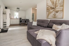 A modern living room with a grey sofa, a dining area with a table and chairs, and a kitchen in the background.