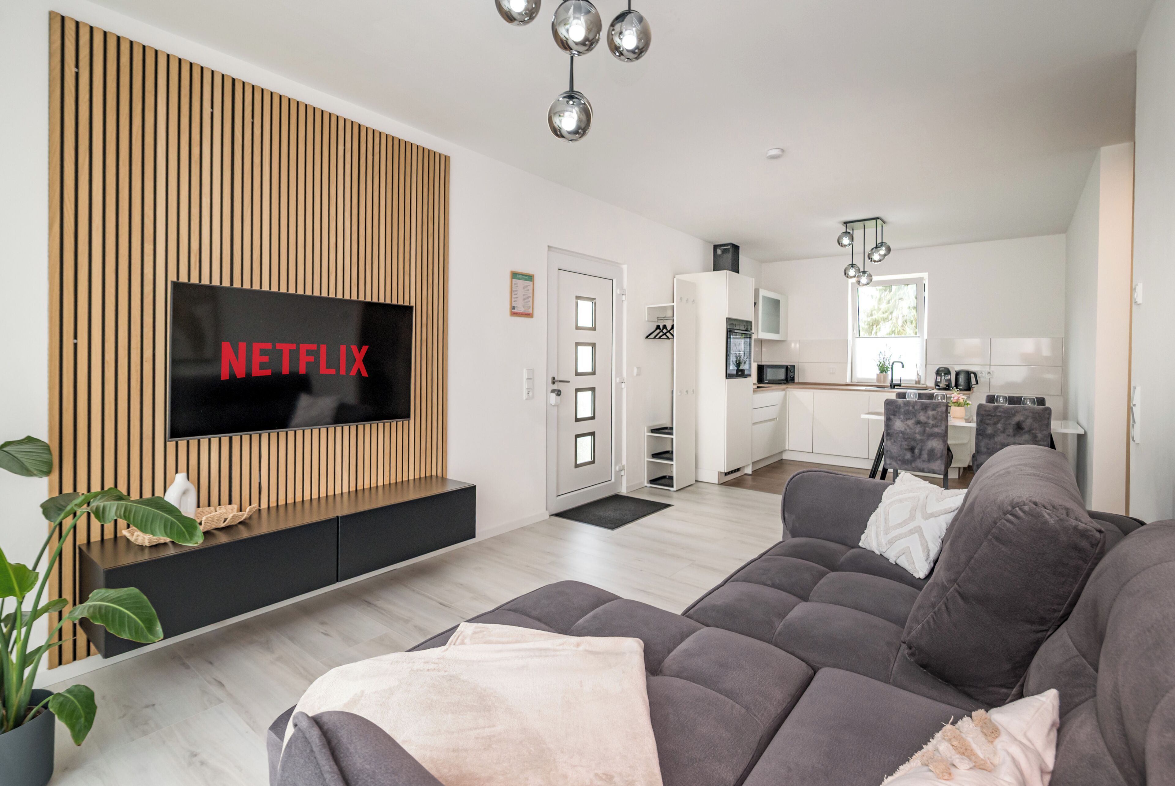 Ein modernes Wohnzimmer mit einem Flachbildfernseher, einer Couch und einem Essbereich, im Hintergrund befindet sich eine Küche.
