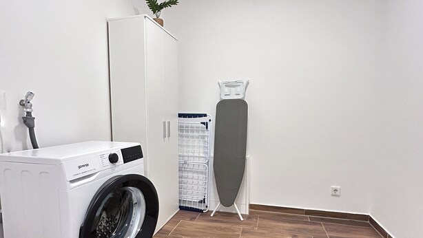 A laundry room with a front-loading washing machine, an ironing board, and a robotic vacuum cleaner.