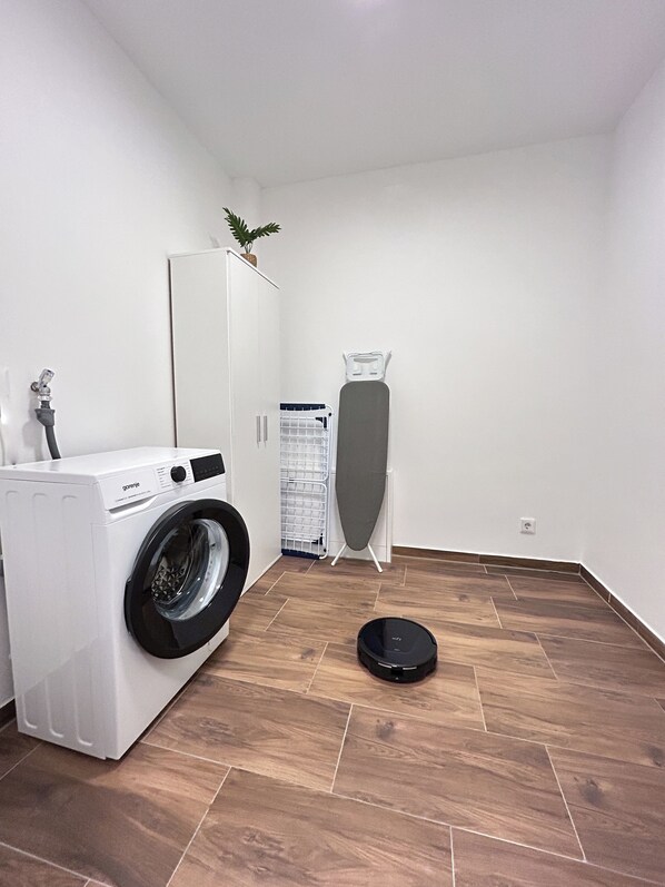 A laundry room with a front-loading washing machine, an ironing board, and a robotic vacuum cleaner.