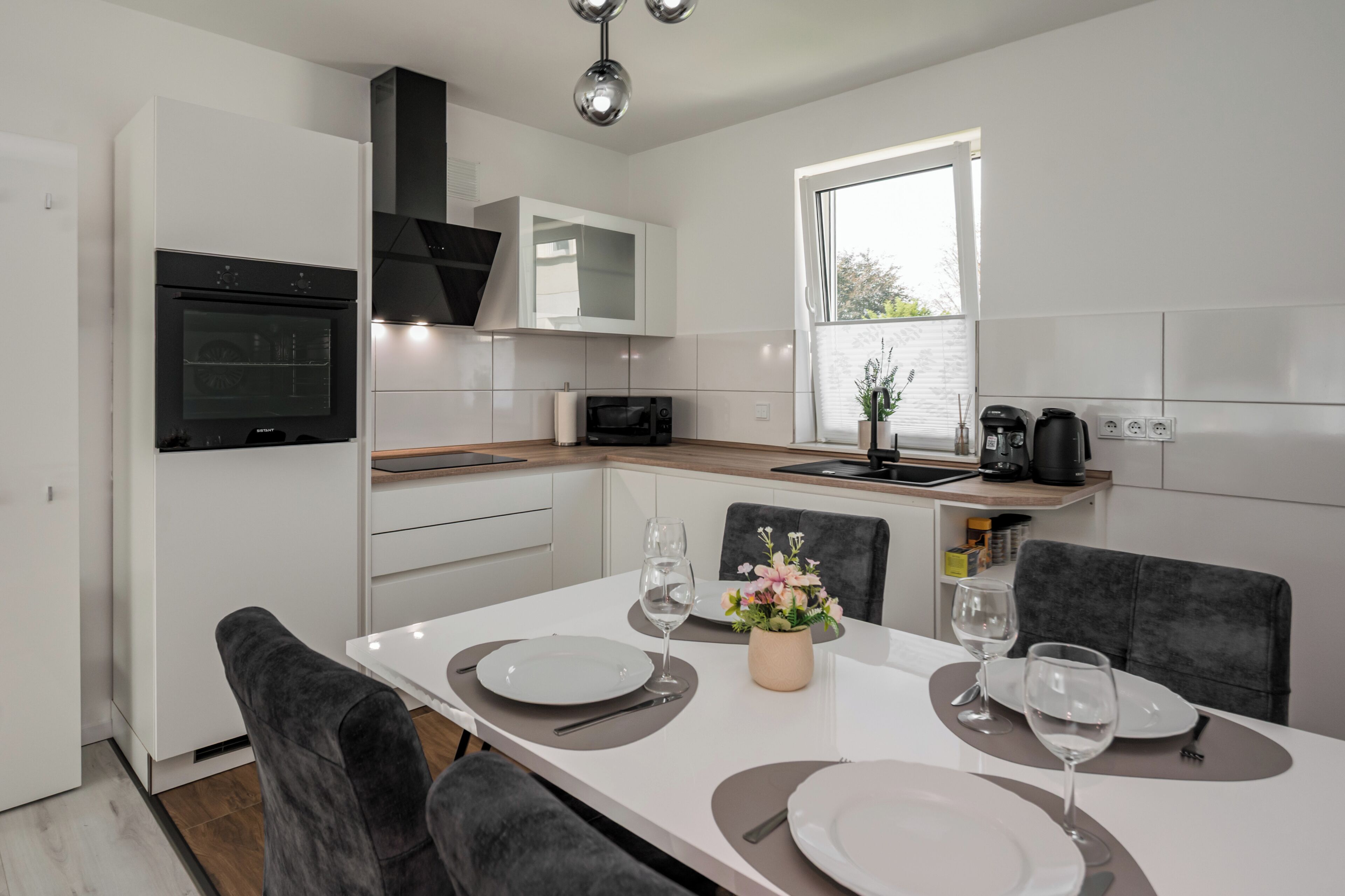 A modern kitchen with white cabinetry, a built-in oven, a microwave, and a coffee maker. There is a dining table set with plates, glasses, and a vase of flowers.