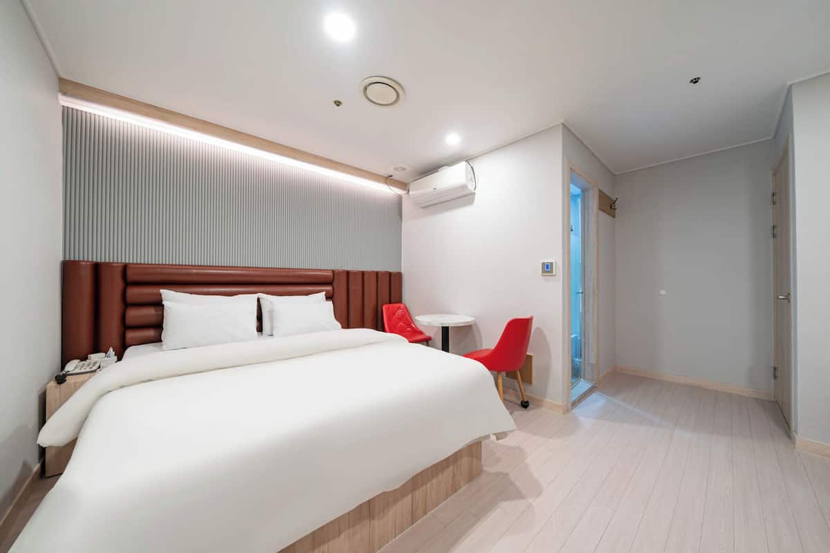 Een moderne hotelkamer met een groot bed, een nachtkastje met een telefoon, een rode stoel, een klein rond tafeltje en een deur naar een andere kamer.