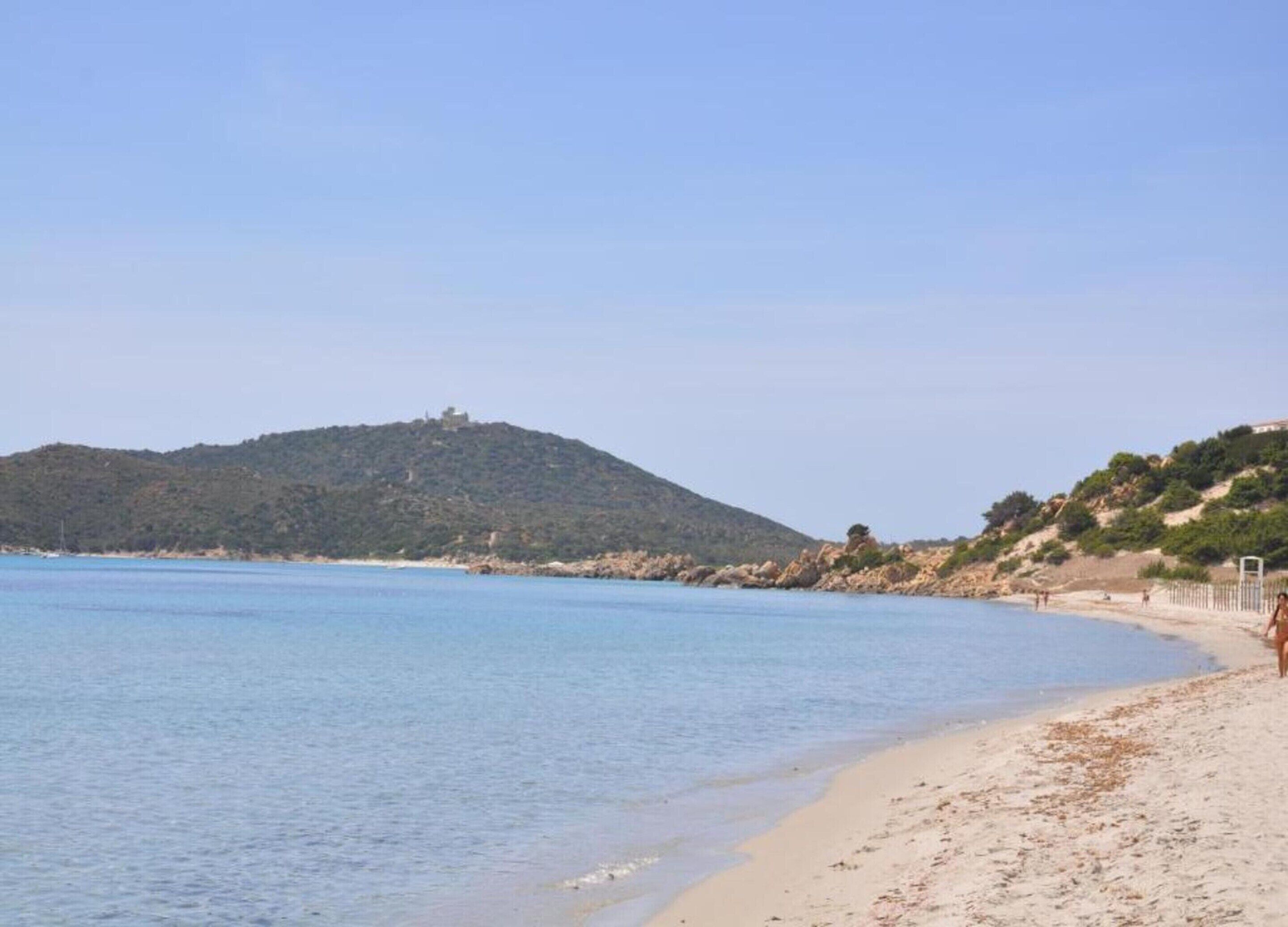A beach with clear blue water, sandy shore, and a hill with vegetation in the background.