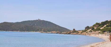 A beach with clear blue water, sandy shore, and a hill with vegetation in the background.