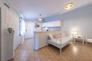 Une pièce de taille modeste comprenant un lit, une petite table avec une lampe, un coin cuisine et une porte donnant sur une autre pièce.