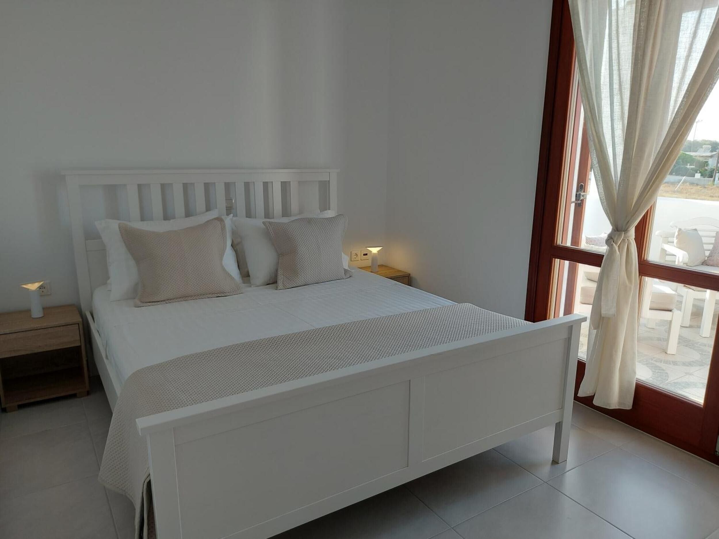 A white bed with a headboard, two bedside tables, and a window with curtains.