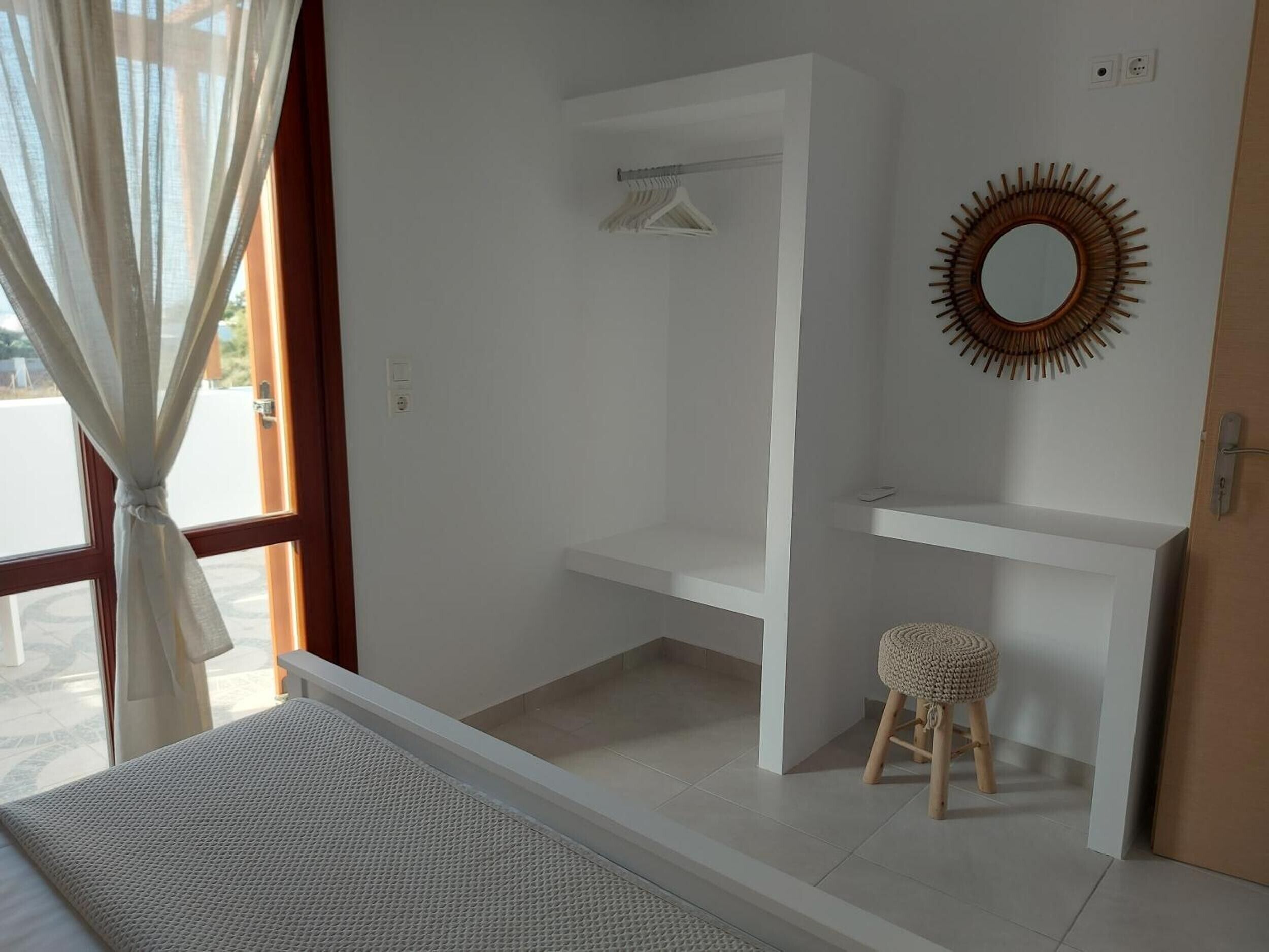 Une pièce comprenant un lit, une armoire blanche, un petit tabouret, un miroir et une fenêtre avec des rideaux.