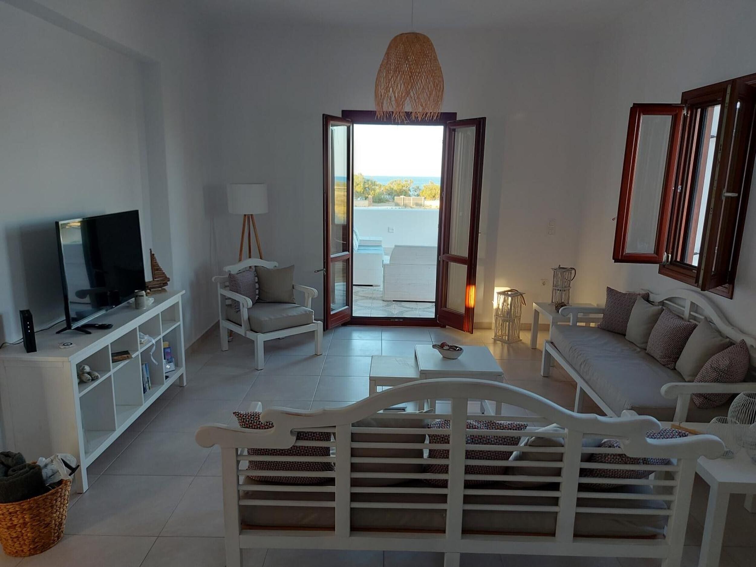 Una sala de estar moderna con televisor, mobiliario blanco, una lámpara de techo de mimbre y vistas a través de un ventanal.