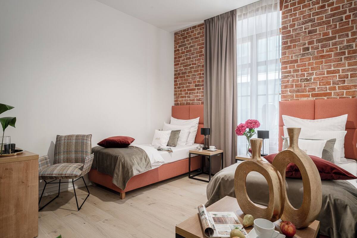 A modern hotel room with a brick accent wall, a large bed, a chair, and a wooden coffee table with a magazine and fruit.