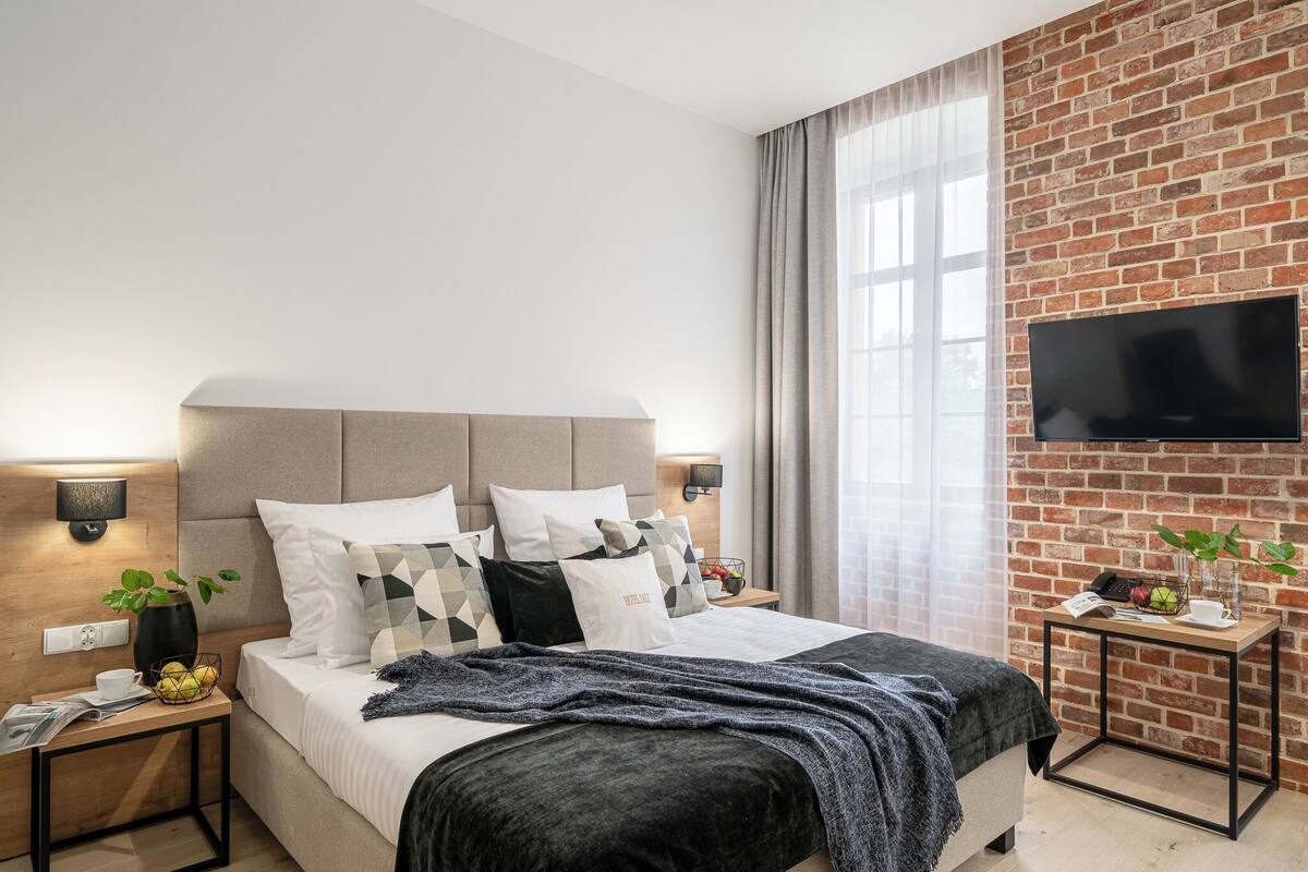 A modern bedroom with a brick wall, a flat-screen TV, a bed with multiple pillows, bedside tables with lamps, and a window with curtains.