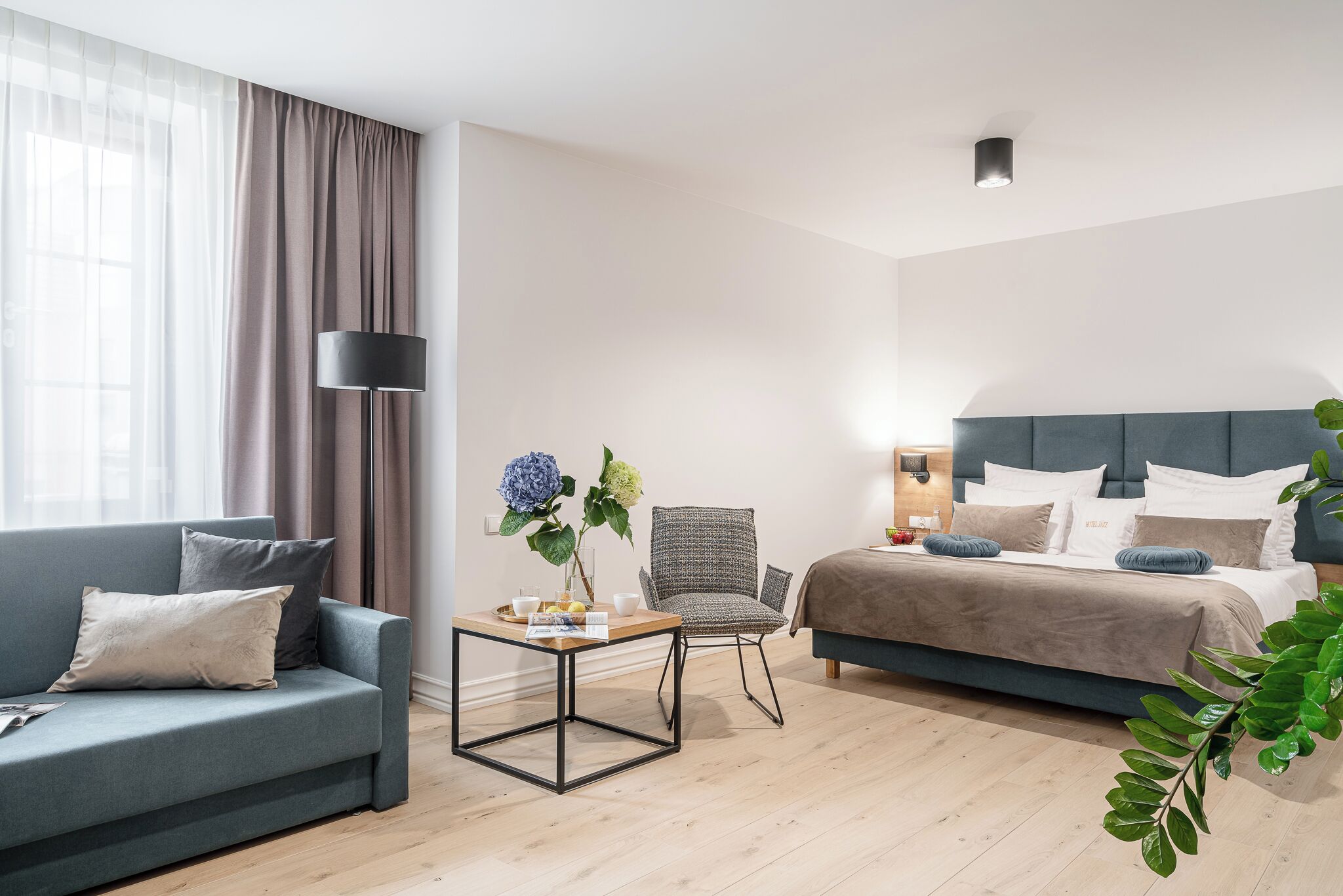 Quarto de hotel moderno com sofá, poltrona, mesa com um vaso, cama com travesseiros e abajur.