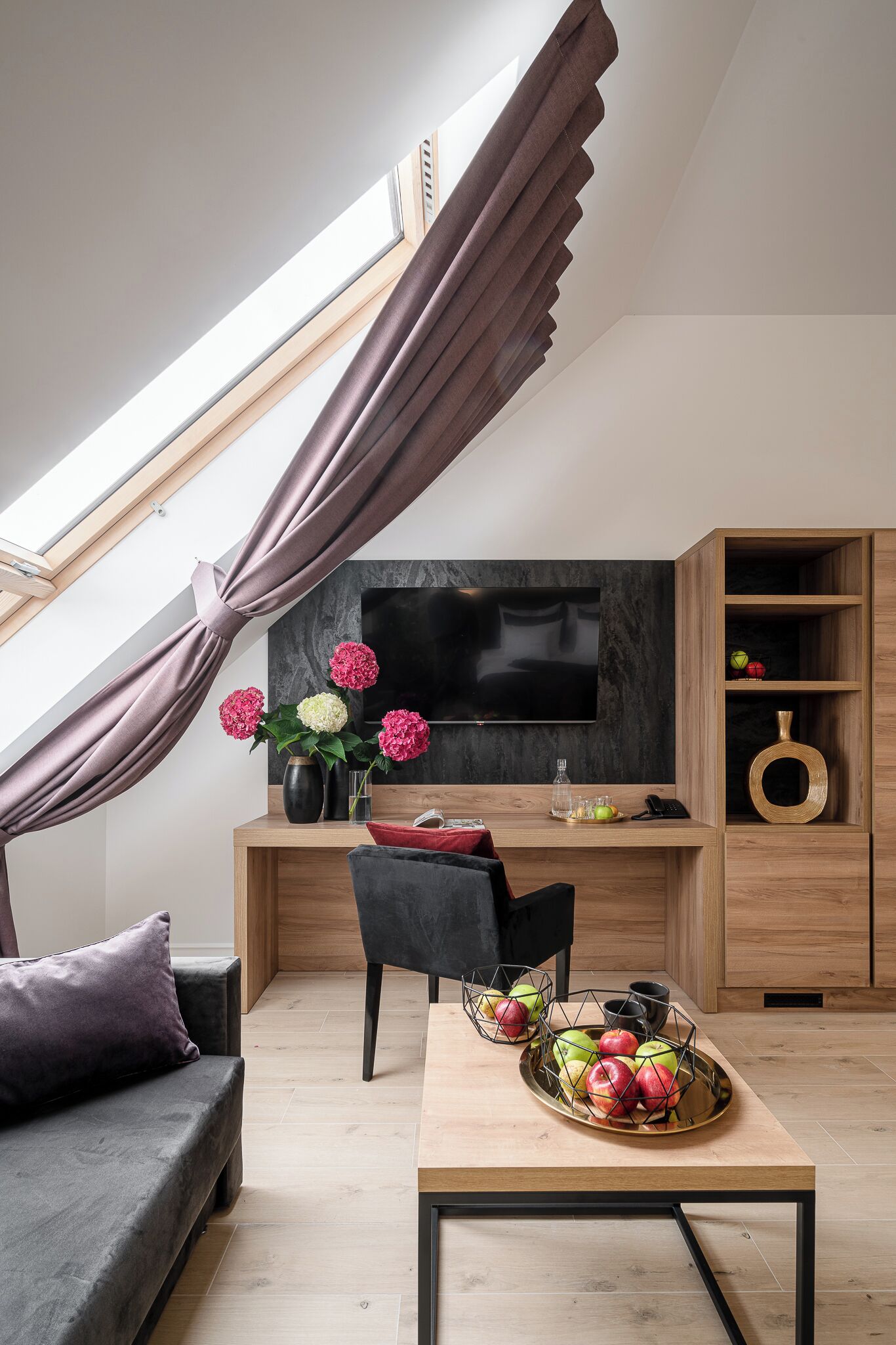 A modern loft bedroom with a skylight, a large flat-screen TV, a wooden desk, and a dining table with fruit.
