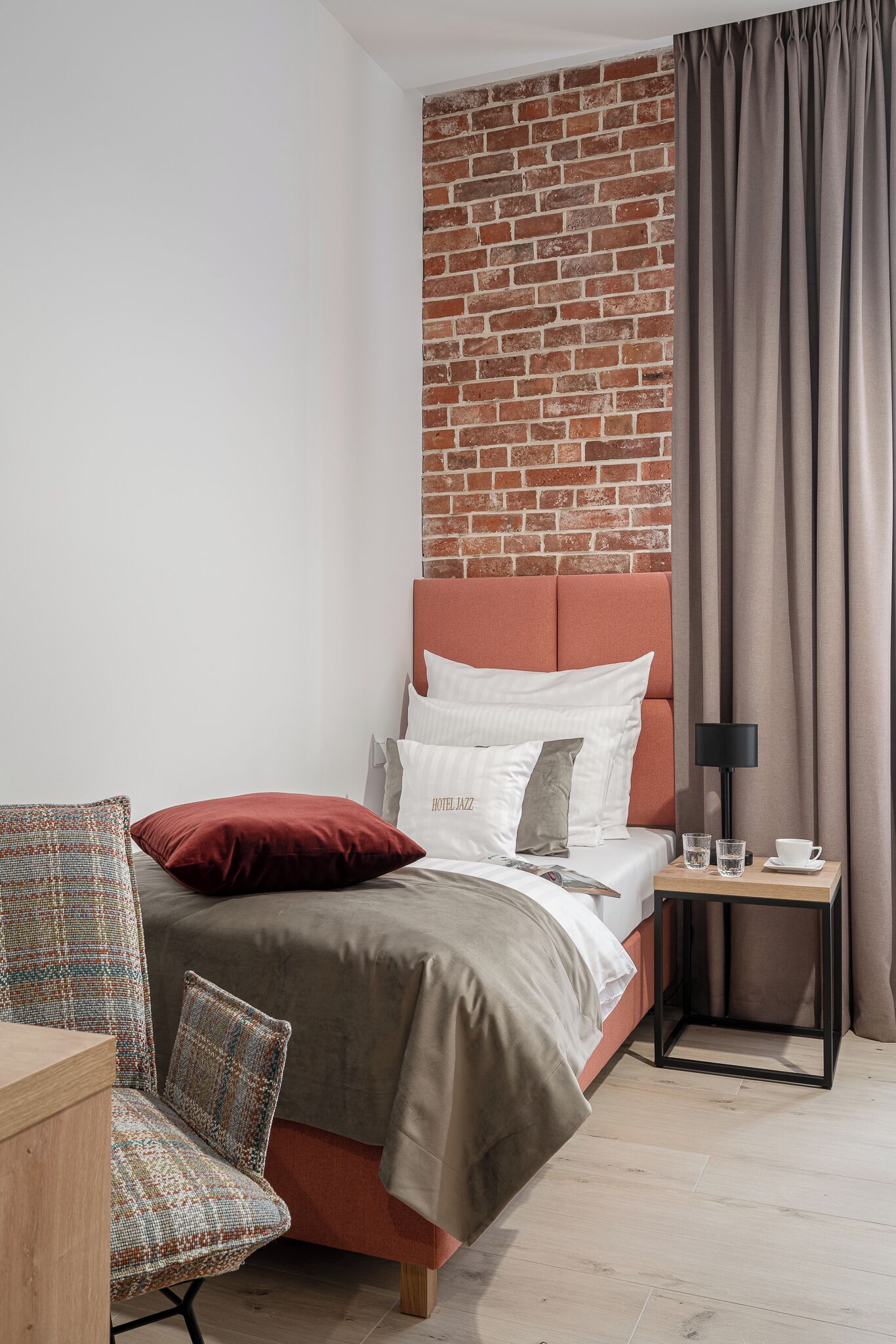 Une chambre d’hôtel moderne avec un mur d’accent en briques, un lit bien fait agrémenté de plusieurs oreillers, une table de chevet avec une lampe et une chaise avec un coussin à motifs.
