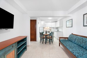 A modern living room with a sofa, a dining table with chairs, a TV mounted on the wall, and a kitchen area in the background.