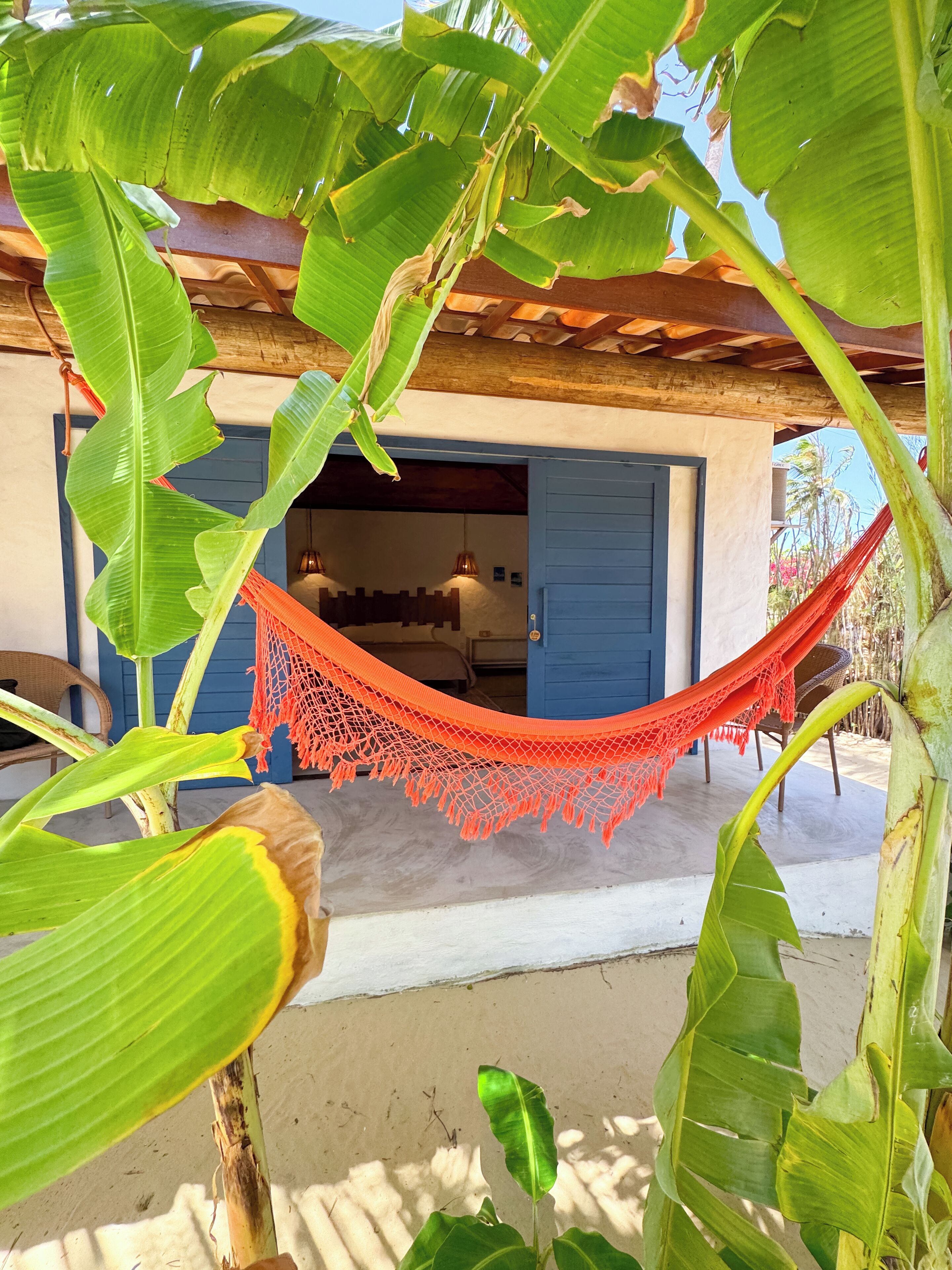 A hammock under banana leaves, a blue door, and a building with a white wall.
