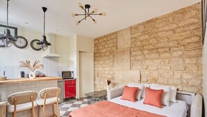 Une chambre avec un mur en pierre, un lit avec une literie orange et un coin cuisine avec un meuble rouge et une horloge.