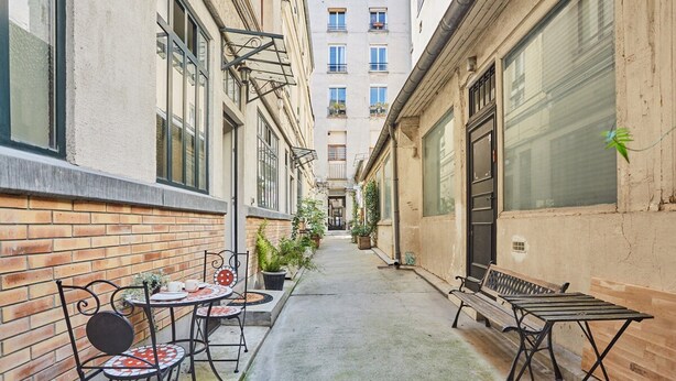 Une ruelle étroite, avec un banc et des tables d’un côté, et un immeuble doté de grandes fenêtres de l’autre.