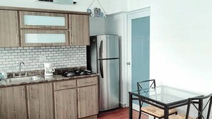 A compact kitchen with wooden cabinets, a stainless steel refrigerator, a glass-top dining table, and a red-tiled floor.
