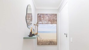 A hallway with a large mirror, a framed beach scene, and a small shelf with decorative items. - Apto no Guarujá com Piscina a Passos da Praia (Guarujá)