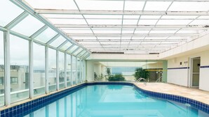 An indoor swimming pool with glass walls and a tiled floor, surrounded by a white interior and a view of the sky. - Apto no Guarujá com Piscina a Passos da Praia (Guarujá)