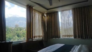A hotel room with a bed, a window with curtains, and a view of mountains. - SOLITUDE (Dharamshala)