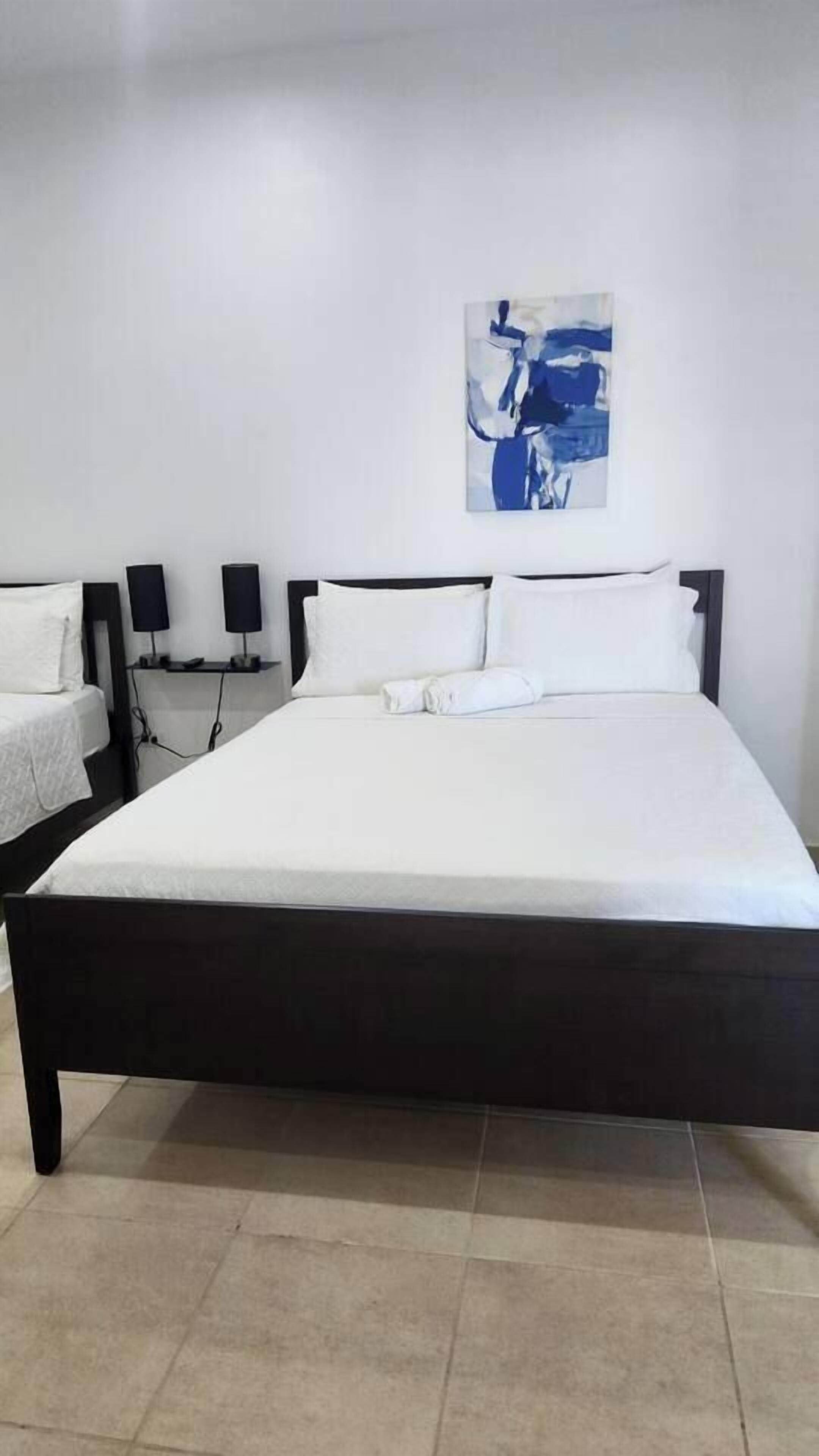 A hotel room with a large bed, two bedside tables with lamps, a white wall, and a blue abstract painting.