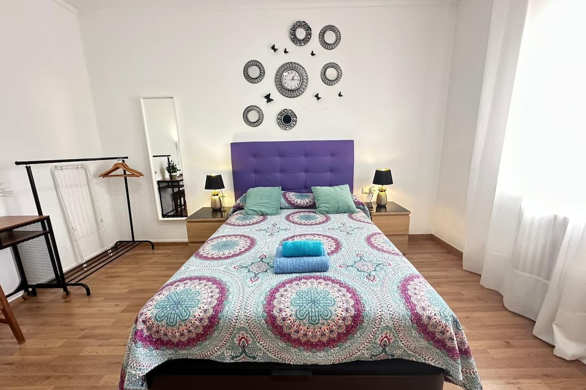 A bedroom with a bed, two bedside tables, a hanging rack, a mirror, and wall decorations.