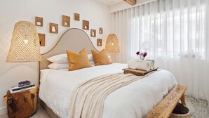 A neatly arranged bedroom with a large bed, two bedside lamps, a bedside table with a book and a decorative item, a window with sheer curtains, and wall-mounted artwork.