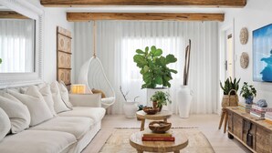 A cozy living room with a white sofa, a hanging chair, a large plant, and a wooden coffee table with books.