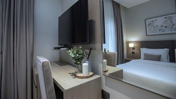A hotel room with a bed, a desk with a mirror, a TV, and a painting on the wall. - Apartment 1010 (Abuja)