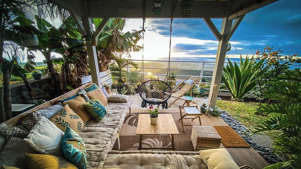 Ein gemütlicher Außenbereich mit gepolstertem Sofa, einer Hängematte und einem Holztisch, umgeben von üppiger Bepflanzung und Blick auf das Meer.