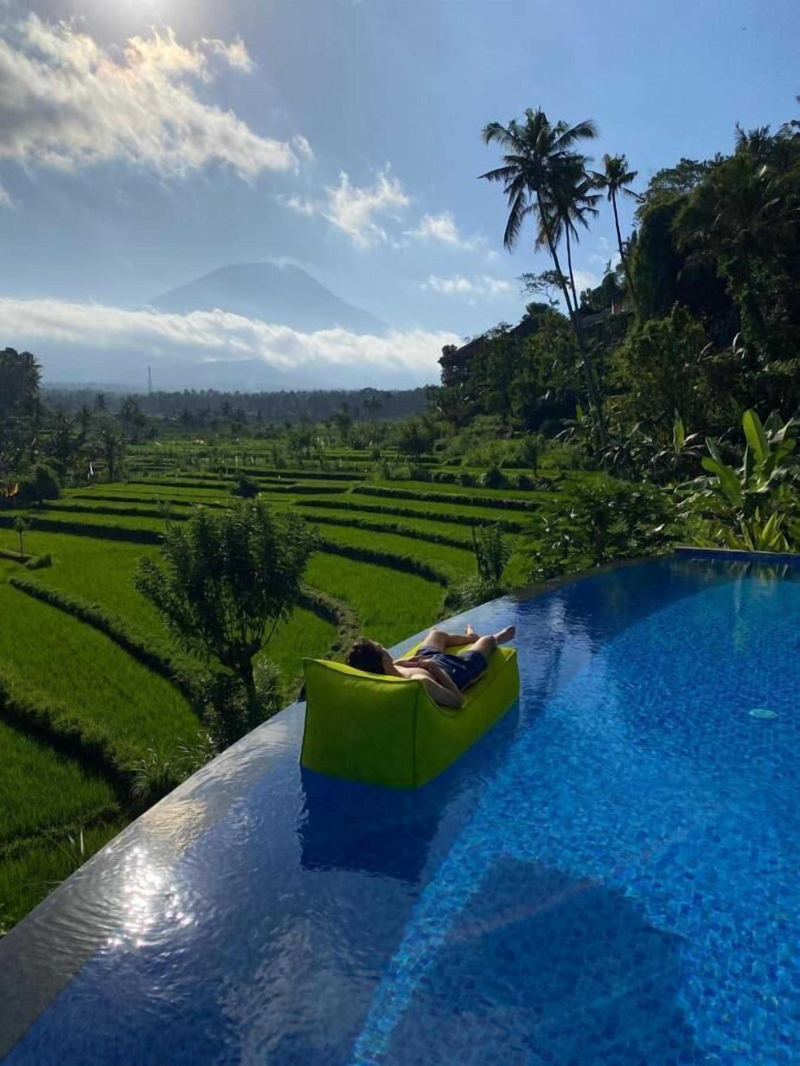 Una persona se relaja en una silla flotante amarilla dentro de una piscina, con vistas a exuberantes campos de arroz verdes y una montaña al fondo.