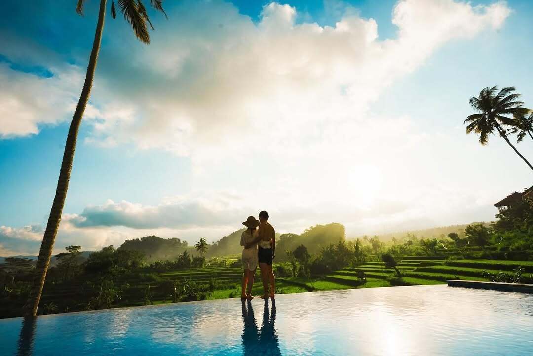 A couple standing by a pool with a view of a lush landscape and palm trees.