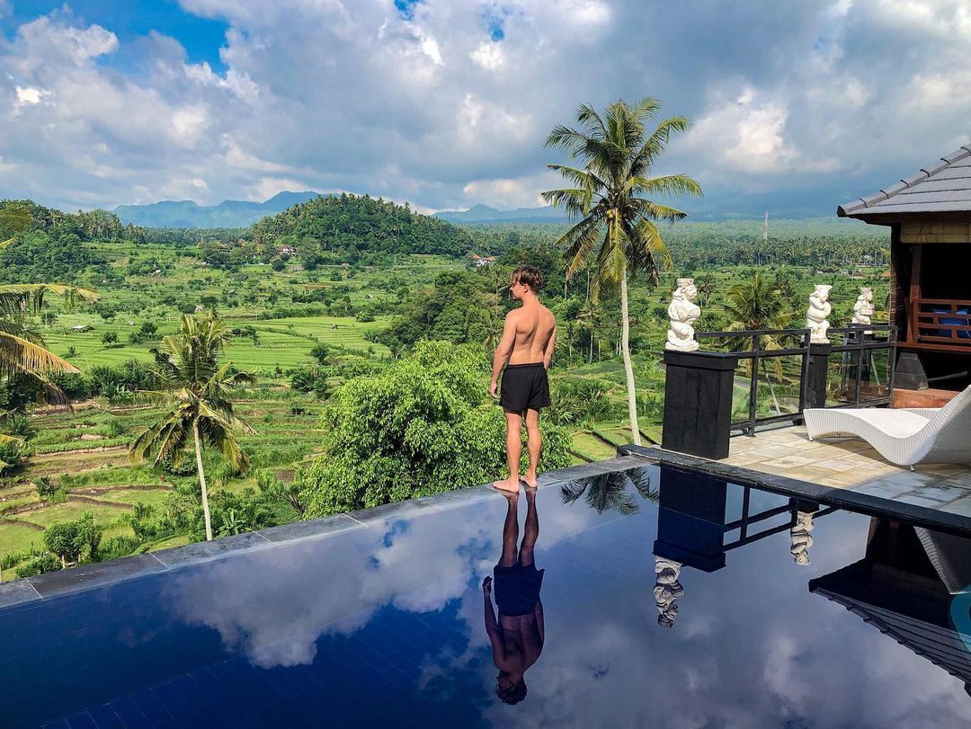 Un homme se tient au bord d’une piscine à débordement, d’où l’on peut admirer un paysage luxuriant et des montagnes en arrière-plan.