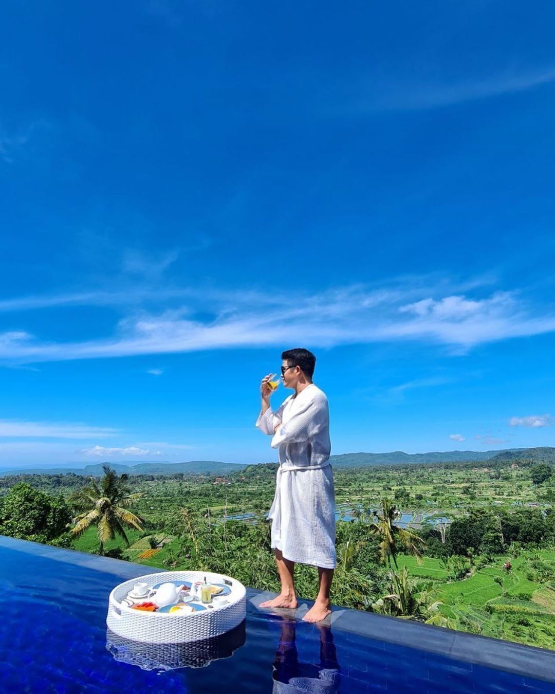 A person in a white bathrobe standing by a pool with a table set for tea and snacks, overlooking a lush green landscape.