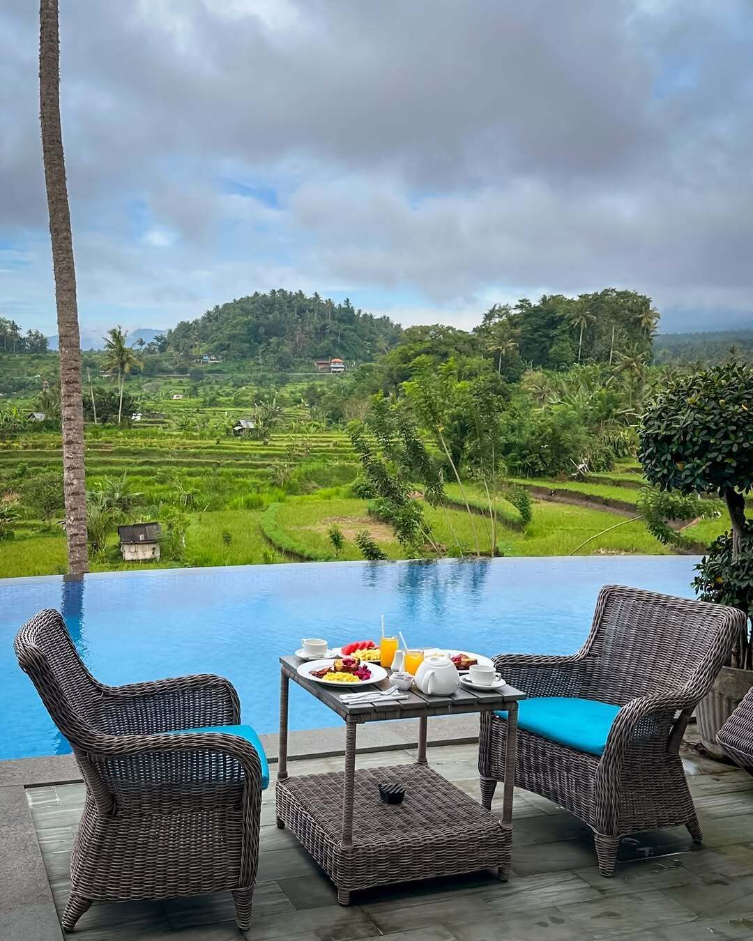 A poolside area with wicker furniture and a table set for breakfast, overlooking lush green fields and a distant hill.