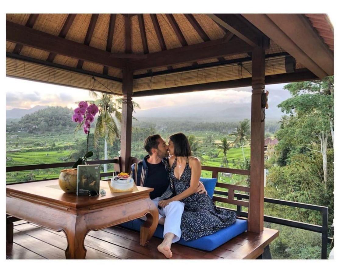 A couple sitting on a wooden bench under a thatched roof, overlooking a lush landscape with a table featuring a cake and a vase of flowers.