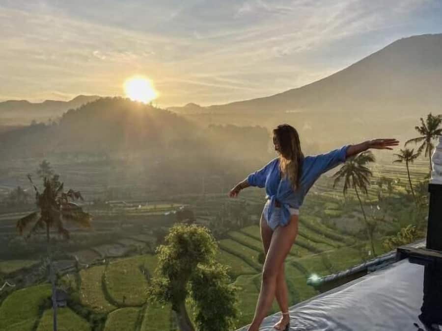 A person standing on a pool edge with a view of a sunset over a hilly landscape.