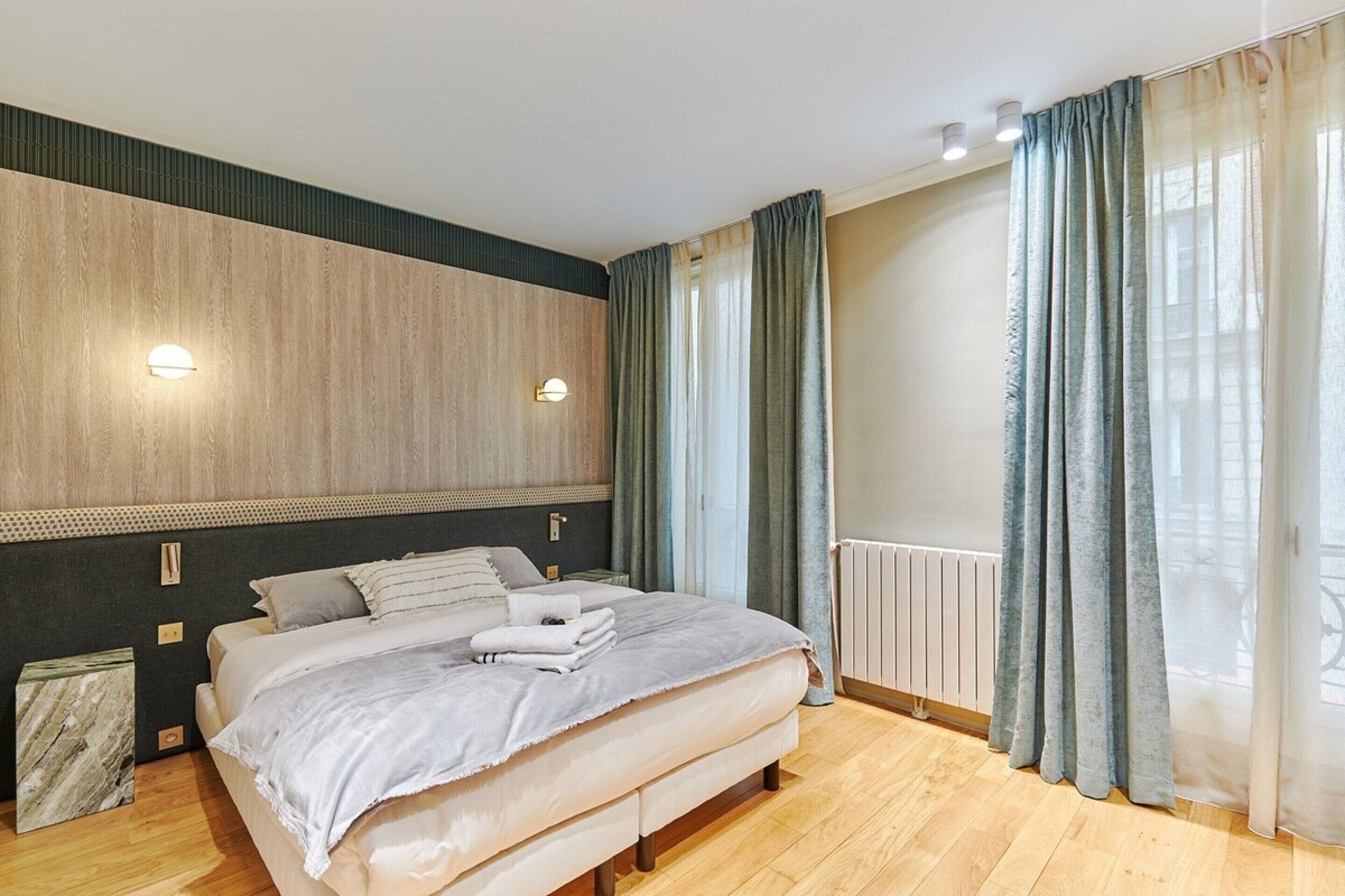 A modern bedroom with a large bed, wooden headboard, and a view of the city through sheer curtains.