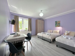 Una habitación con dos camas, un escritorio, una silla, un ventanal con cortinas, un radiador y un cuadro en la pared.