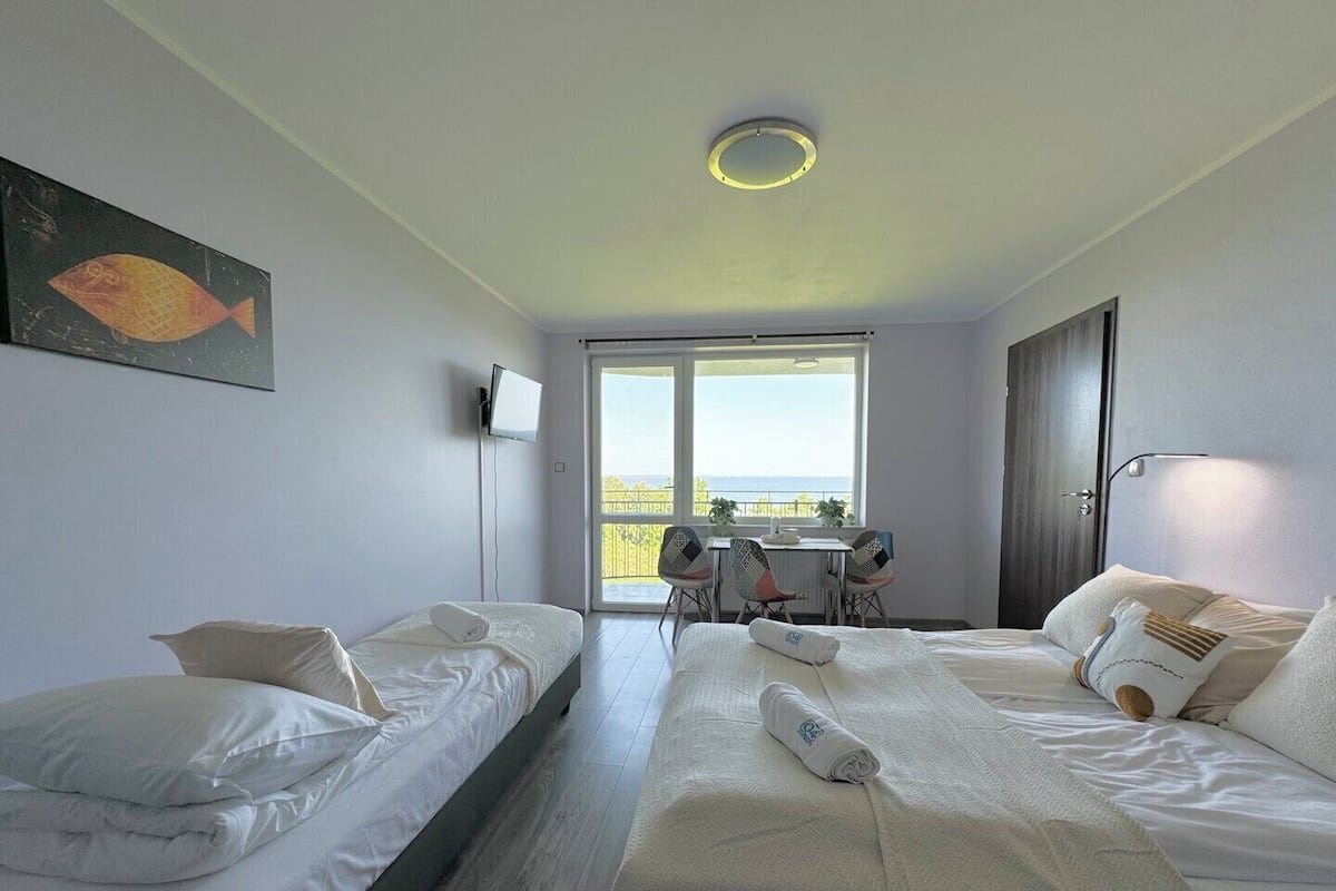 A hotel room with two single beds, a large bed, a TV, a balcony with a table and chairs, and a view of the sea.